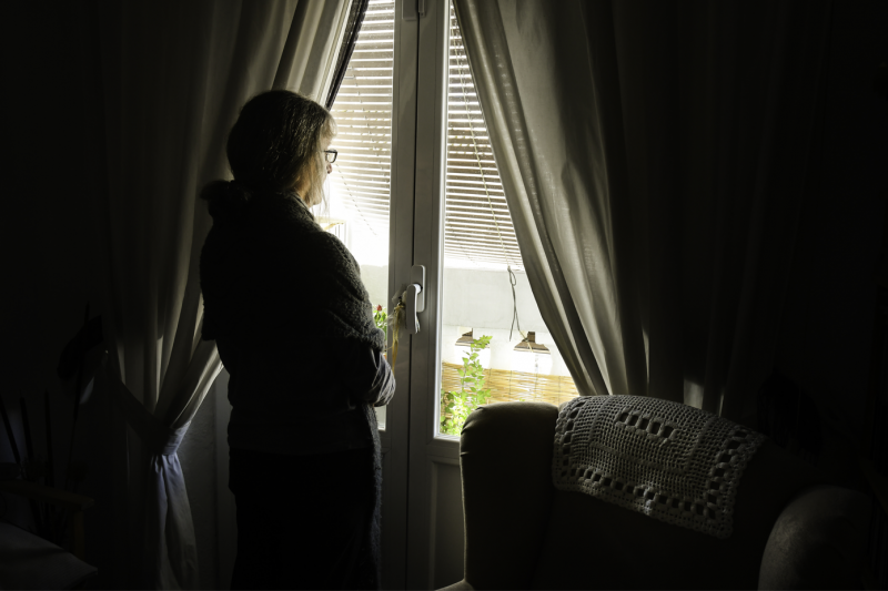 Mujer adulta, confinada por la pandemia, mira hacia la calle a través de la ventana de su casa