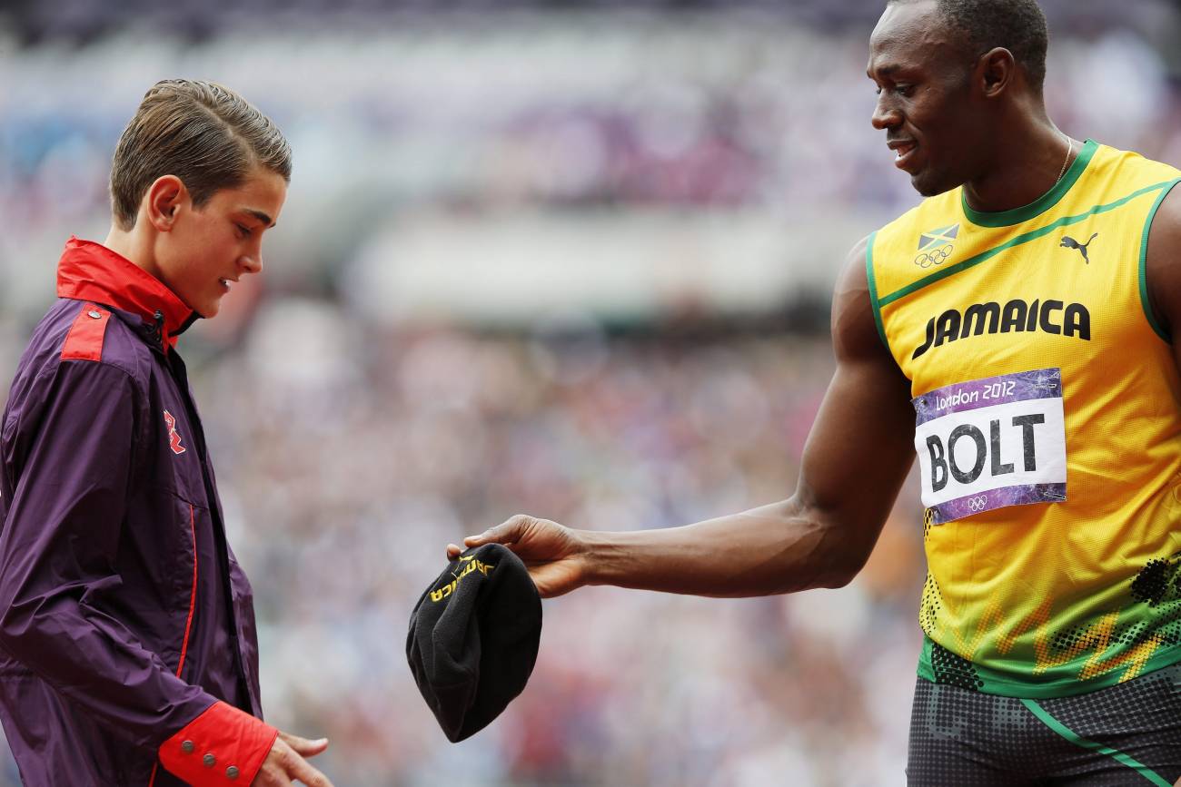 El atleta jamaicano Usain Bolt (dcha) le entrega su gorra a un voluntario antes del comienzo de las mangas clasificatorias de los 200m masculino en la competición de atletismo de los Juegos de Londres